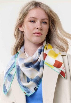 Blonde Frau mit blauen Augen, die einen cremefarbenen Mantel, ein blaues Oberteil und einen mehrfarbigen karierten Schal mit türkisfarbener Umrandung trägt und vor einem einfarbigen Hintergrund nach vorne blickt.