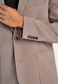 Beżowa marynarka o gładkiej fakturze, z pojedynczą kieszenią na piersi i czterema czarnymi guzikami na mankiecie. Charakteryzuje się dopasowanym krojem i minimalistycznym designem.