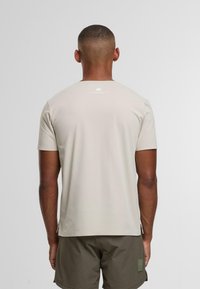 Uomo con capelli corti in piedi di spalle, indossa una camicia beige a maniche corte e pantaloni corti verde oliva, su uno sfondo chiaro e uniforme.