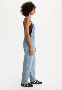 Ανοιχτό μπλε τζιν ολόσωμες φόρμες με φαρδιά πόδια, ρυθμιζόμενους ιμάντες και εμπρόσθιες τσέπες, φορεμένες πάνω από ένα ναυτικό μπλε και κίτρινο αμάνικο μπλουζάκι; συνδυασμένες με μαύρα αθλητικά παπούτσια.