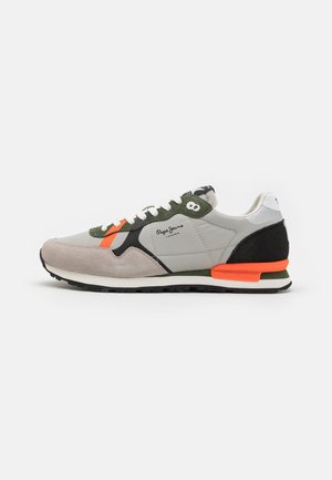 Zapatilla gris y negra con acentos naranja y verde, mezcla de texturas suaves y de ante, puntera redonda, cordones blancos, suela negra, logotipo de la marca visible.