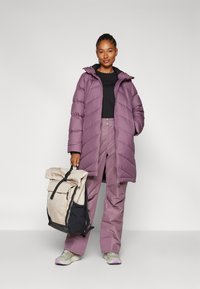 Lila puffjacka med huva, svart topp och matchande lila byxor. Beigefärgad och svart ryggsäck med rulltop. Sneakers med grå och lila detaljer.