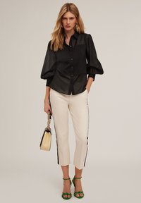 Camicia nera con maniche a sbuffo, abbinata a pantaloni crema sartoriali con strisce nere laterali e sandali verdi con cinturini.
