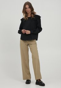 Schwarze Bluse mit einem strukturierten, gewellten Kragen, langen Ärmeln, lockerer Passform, kombiniert mit beigen weiten Hosen und schwarzen Slippern.