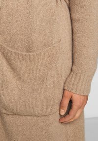 Beżowy sweter dzianinowy z prążkowanymi mankietami i dołem, z dużą kieszenią z przodu i miękką, teksturowaną powierzchnią. Zbliżenie na rękę spoczywającą na tkaninie.