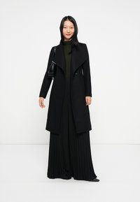 Casaco preto em mistura de lã com colarinho drapeado, botões duplos e um vestido maxi preto pregueado por baixo. Design minimalista, sem padrões.
