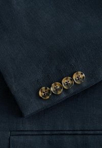 Manche de costume bleu marine avec quatre boutons en écaille de tortue marron disposés en ligne au-dessus d'une poche à rabat.