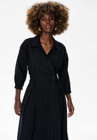 Czarna sukienka kopertowa z rękawami 3/4, kołnierzykiem, zapięciem na guziki w talii oraz zakładkami na spódnicy dla zwiększenia objętości.