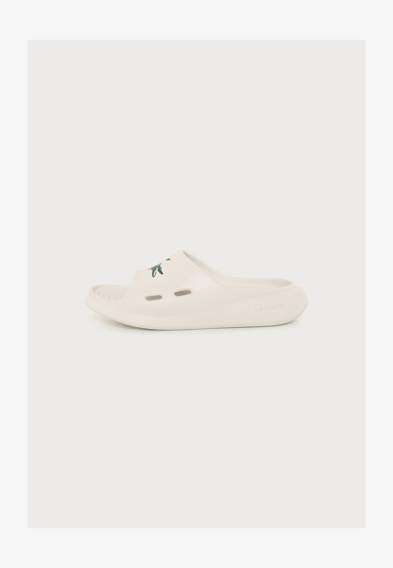 Λευκό σαγιονάρι Lacoste με τρύπες αερισμού και ανάγλυφο πράσινο λογότυπο κροκόδειλου στο πάνω λουράκι, σε μονόχρωμο φόντο.
