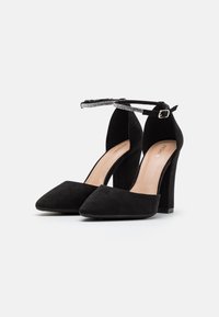 Chaussures noires en daim à talons hauts avec des bouts pointus et des brides de cheville ornées de petits strass, sur une surface blanche.