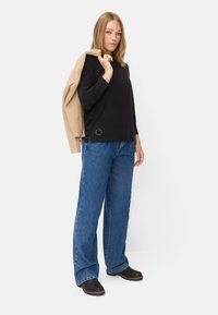 Schwarzes Langarmoberteil, blaue weite Jeans und schwarze Clogs; brauner Strickpullover über die Schulter gelegt. Auffälliges Logodetail am Saum.