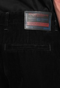 Μαύρο τζιν από κορντόρι με ομαλή, ριγωτή υφή. Διαθέτει δερμάτινη ετικέτα με κεντητικό μοτίβο που αναγράφει το όνομα της μάρκας και δήλωση ποιότητας.