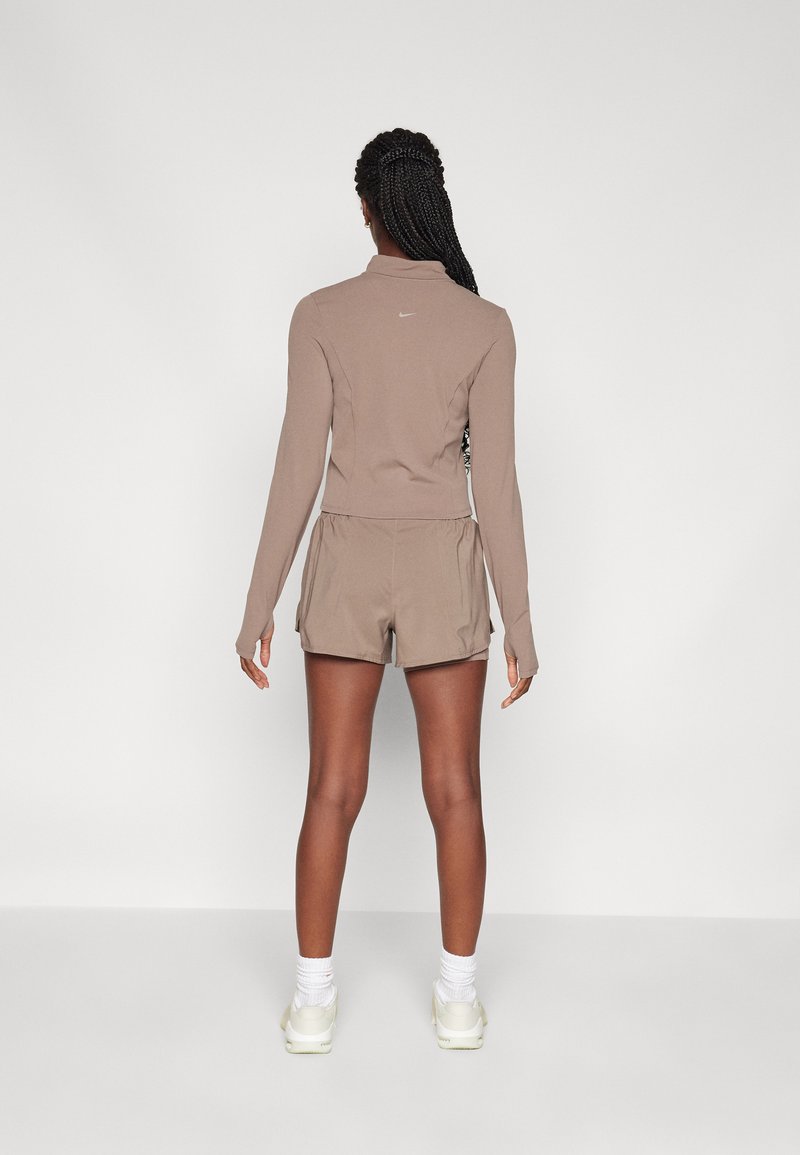 Maglietta a maniche lunghe, aderente, di colore marrone chiaro con colletto alto e logo bianco sul retro, abbinata a pantaloni corti beige larghi e calze bianche.