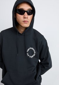 Sweat-shirt à capuche noir avec un logo circulaire blanc comportant le texte "new balance". Le tissu est doux, avec une poche kangourou et un cordon de serrage pour la capuche.