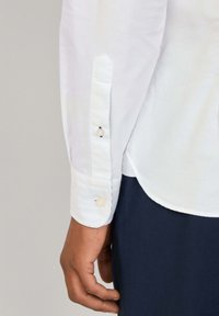 Camisa blanca con puño abotonado, que presenta un diseño de manga puntiaguda y una textura sutil. Tela azul marino visible debajo.