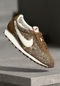 Bruine gestructureerde sneaker met een witte Swoosh, bovenwerk van textiel, en bruine veters met een rubberen zool en gripnoppen.