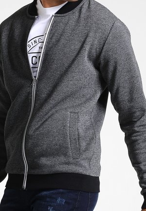 Mies, jolla on harmaa vetoketjullinen takki mustalla kauluksella ja hihansuuilla, valkoisen graafisen T-paidan ja tummansinisten farkkujen päällä.