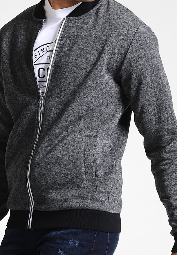 Homme portant une veste grise à fermeture éclair avec col et poignets noirs, par-dessus un T-shirt blanc à motif et un jean bleu foncé.