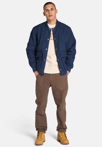 Marinblå quiltad bomberjacka med tryckknappar, två framfickor, beige sweatshirt under, bruna cargobyxor och gula snörboots.