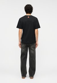 Μαύρο βαμβακερό μπλουζάκι με στρογγυλή λαιμόκοψη, κοντά μανίκια και μικρό κόκκινο λογότυπο στο πάνω μέρος της πλάτης. Φορεμένο με άνετο σκούρο τζιν τζιν.