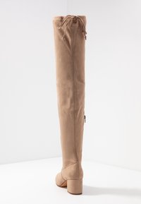 Botas de ante beige hasta la rodilla con punta redonda, tacón grueso y detalle de cordones en la parte superior. Cremallera lateral para facilitar su uso.