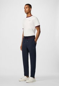 Witte t-shirt met een kastanjebruine ribbelkraag, gecombineerd met marineblauwe trainingsbroeken met een trekkoord in de taille en een logo-detail, en witte sneakers.