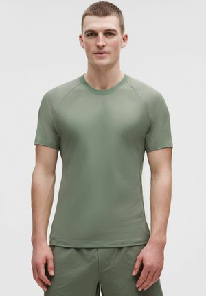 Giovane uomo con indosso una maglietta aderente a maniche corte verde oliva e pantaloncini coordinati, in piedi contro uno sfondo chiaro e semplice.