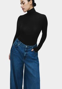 Vrouw met donker haar in een zwarte coltrui en wijde blauwe jeans poseert met één hand in de zak tegen een effen achtergrond.