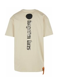 Béžové bavlněné tričko s vertikálním černým nápisem "zapomenutí fanoušci" na zadní straně a kulatou černou nášivkou nad ním. Oranžová nálepka na boku.