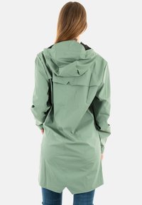 Persoon in een lichtgroene regenjas met capuchon en lange mouwen, met de rug naar de camera gekeerd, in combinatie met blauwe jeans tegen een effen achtergrond.