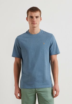 Jonge man die een blauw T-shirt met korte mouwen draagt met een klein geel logo op de borst en lichtgroene broek, staand tegen een witte achtergrond.