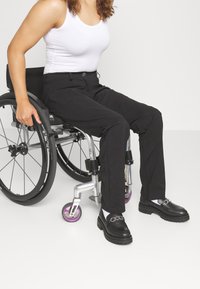 Black pants with a slim fit and pockets, paired with black loafers featuring metallic chain accents and white ankle socks, worn in a wheelchair.