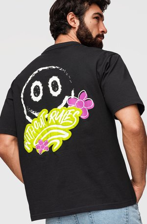 Homme portant un t-shirt noir avec un grand visage souriant blanc, texte jaune fluo "WITHOUT RULES" et motifs floraux roses dans le dos.