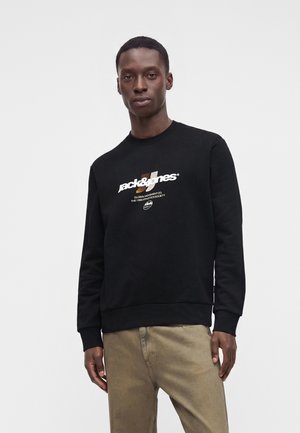 Homme portant un sweat-shirt noir Jack & Jones et un pantalon beige, debout devant un fond clair uni, regardant droit devant.