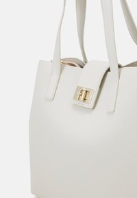 Sac en cuir texturé blanc avec quincaillerie dorée, présentant une sangle de fermeture proéminente et deux poignées robustes pour l'épaule.