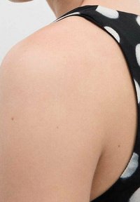 Tissu noir avec des pois blancs ; présente un design sans manches et une épaule creusée. Texture lisse et silhouette ajustée.