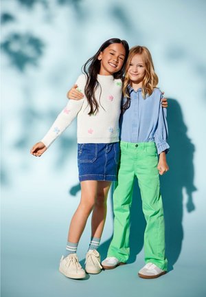Deux enfants se tiennent côte à côte. L'un porte un pull blanc côtelé avec des accents floraux colorés et une jupe en jean, tandis que l'autre porte une chemise bleu clair et un pantalon vert.