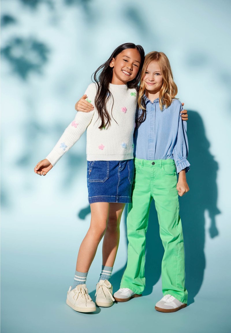 Divi bērni stāv blakus. Vienam ir balts ribots džemperis ar krāsainiem ziedu akcentiem un džinsa svārki, bet otram gaiši zila krekls un zaļas bikses.