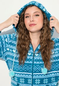 Tuta in pile turchese con cappuccio, caratterizzata da motivi geometrici bianchi e pon-pon sui lacci. Chiusura frontale con zip.