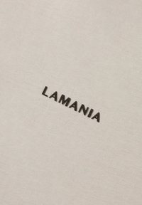 Světle béžová tkanina s jemnou strukturou, na které je vyšitý nápis "LAMANIA" černou barvou. Text je umístěn ve středu.