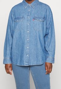 Camicia di jeans azzurro chiaro con maniche lunghe, chiusura a bottoni frontale e due tasche sul petto. Presenta un colletto classico e una vestibilità rilassata.