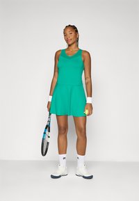 Idrettsutøver i en grønn tennisdrakt holder et racket og en tennisball, iført sportsko og håndleddsbånd, og smiler selvsikkert.