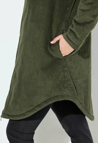 Groene corduroy jas met een gebogen zoom en zijzakken, met ribbelstructuur en lange mouwen, gecombineerd met zwarte broeken.