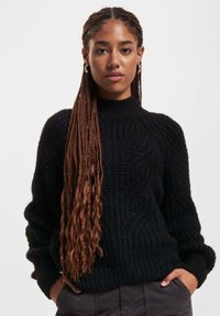 Schwarzer Kabelstrickpullover mit hohem Kragen und langen Ärmeln, der ein strukturiertes Muster und eine lockere Passform aufweist. Trägt man mit den Händen in den Taschen.