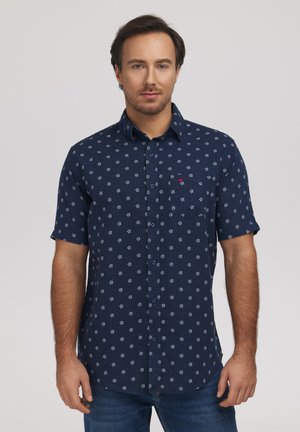 Mężczyzna w granatowej koszuli z krótkim rękawem zapinanej na guziki z drobnym białym kwiatowym wzorem, stojący na tle jasnego, jednolitego tła.