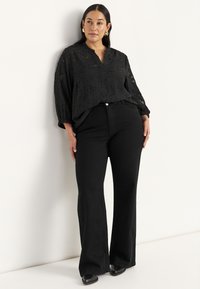Sort, tekstureret bluse med V-hals, lange ærmer og elastiske manchetter, parret med høj talje, flagrende sorte jeans og sorte sko.