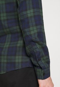 Camisa de franela a cuadros verdes y azul marino con mangas largas, que presenta botones negros y un bajo recto. La tela tiene una textura suave.