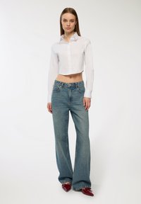 Camicia bianca corta con bottoni e maniche lunghe, abbinata a jeans chiari a zampa d'elefante e tacchi rossi lucidi. Tessuto morbido e design informale.