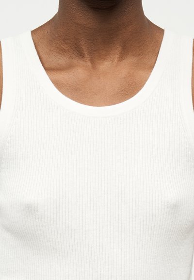 Κοντινό πλάνο προσώπου ατόμου που φοράει λευκή ριμπ μπλούζα χωρίς μανίκια, με εμφανή τον λαιμό, τους ώμους και το πάνω μέρος του στήθους.