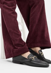 Mocassins en cuir noir avec un accent en garniture dorée, accompagnés de pantalons en velours côtelé bordeaux présentant une texture côtelée verticale.